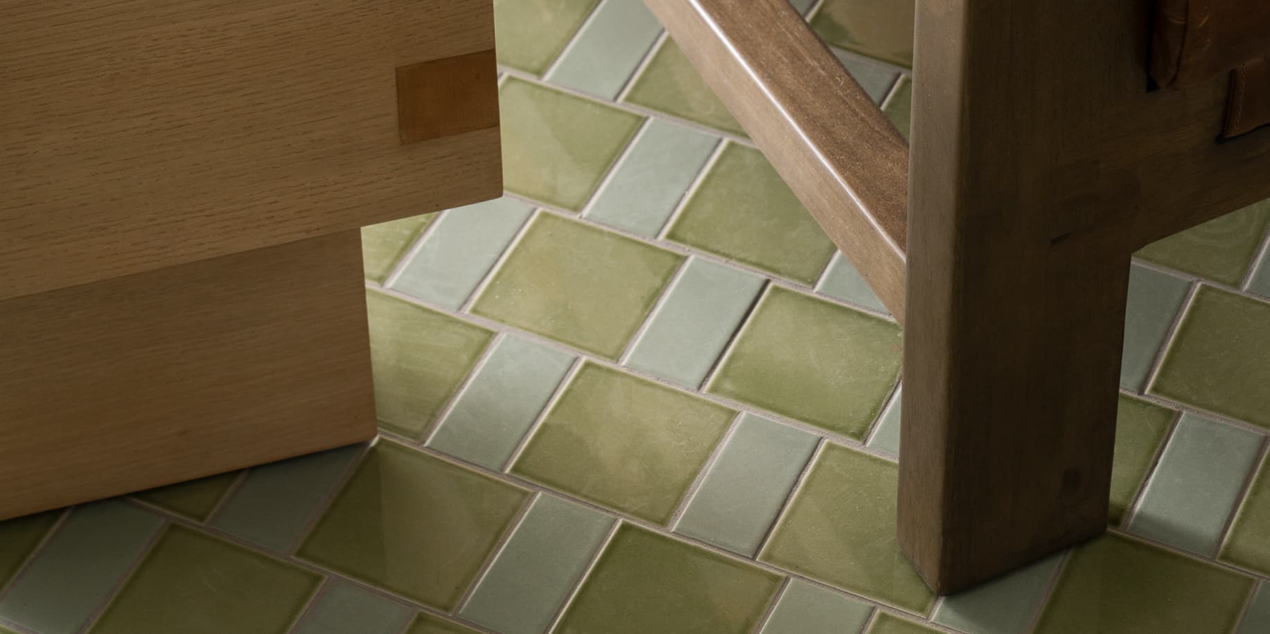 Close-up of sage green rectangular tiles arranged in a staggered pattern on the floor, with wooden furniture legs partially visible in the frame.