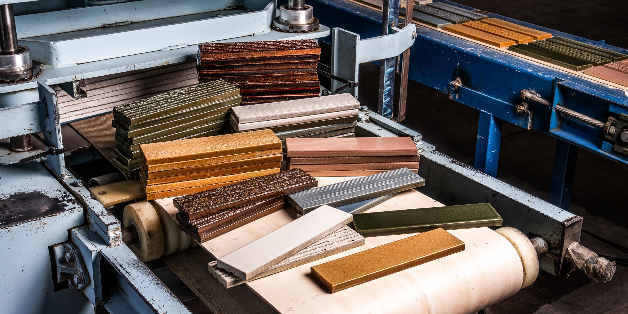 Rectangular ceramic tiles in various colors and finishes are stacked and scattered on the conveyor belt of an industrial tile manufacturing machine.